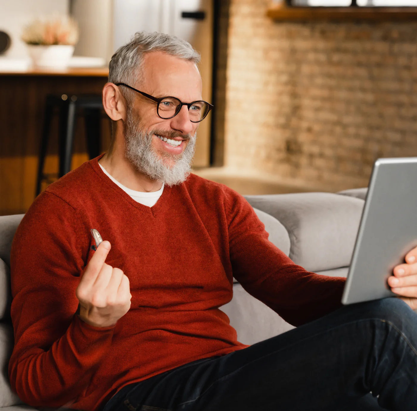 A middle-aged man with salt-and-pepper hair and glasses, wearing a red sweater, is seated comfortably on a gray couch. He smiles engagingly while holding a stylus and interacting with a tablet. The setting is a cozy living room with a warm and inviting decor.