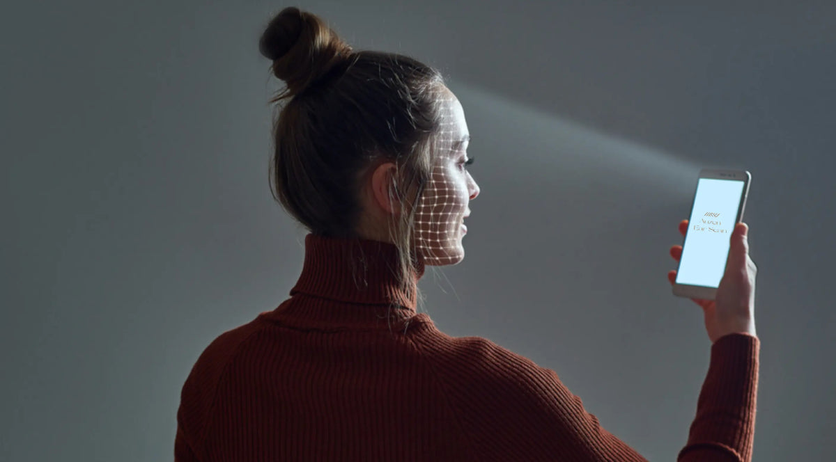 A young woman in a dark red turtleneck uses her smartphone, which displays a facial recognition grid on her face. The setting is simple with soft lighting and a gray background, emphasizing modern technology.