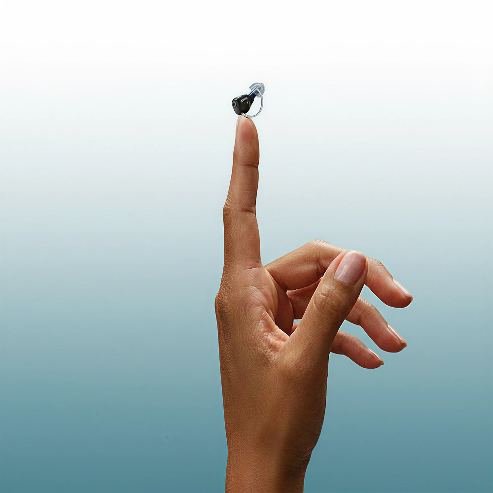 A hand with fingers gracefully extended holds Oticon’s Zeal hearing aid balanced on the tip of the index finger against a gradient blue background.