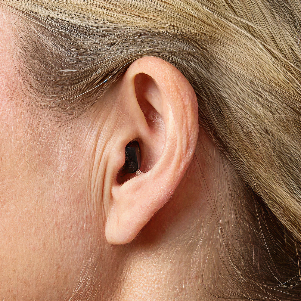 Close-up of a person’s ear wearing the Oticon Zeal™ 1, a small black in-ear hearing aid, fitted discreetly in the ear canal. The individual’s skin and blonde hair are visible.