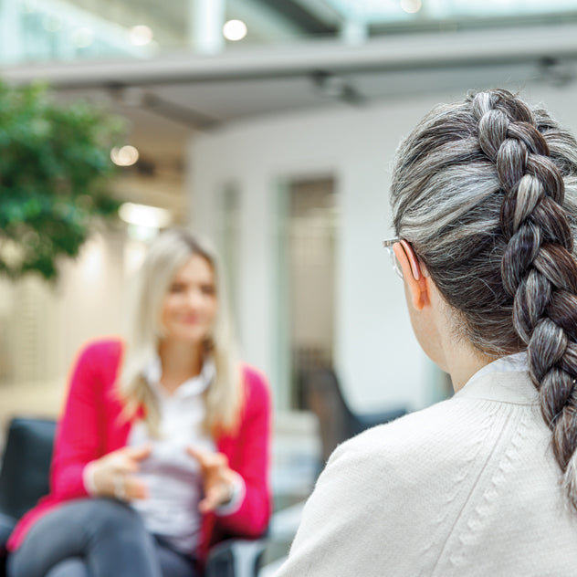 A woman with grey hair in a braid is seen from the back, wearing a Phonak Slim 90 hearing aid, having a conversation with another woman in a red blazer in a modern office setting.