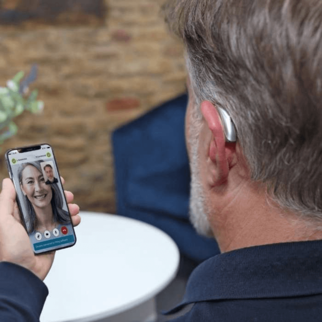 An elderly man wearing Phonak Lumity L90-RT and L50-RT hearing aids is holding a smartphone while on a video call. The screen shows a smiling woman on the other end of the call. The man is seated in a cozy indoor setting with a brick wall and a small table in the background.